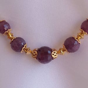 Op de afbeelding: Close-up van een ketting met dieppaarse, facetgeslepen kralen, afgewisseld met goudkleurig filigraan en kleine, goudkleurige accentkralen. De ketting is geplaatst tegen een zachte, gebroken witte achtergrond.