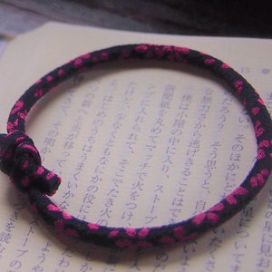 Op de afbeelding: Een zwart-roze armband met een geknoopte sluiting. De armband is gemaakt van geweven materiaal en ligt op een open boek met Japanse tekst.
