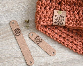 Op de afbeelding: Een oranje haakwerk met een "HAND MADE" leren label en twee extra leren labels. De labels hebben een logo van een bol wol. Twee messing klinknagels zijn ook zichtbaar. De items liggen op een licht houten oppervlak.