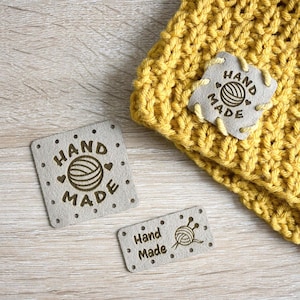 May include: Close-up of three grey leather tags with the words "Hand Made" and a yarn ball design. One tag is attached to a yellow knitted item. The tags have small holes around the edges.