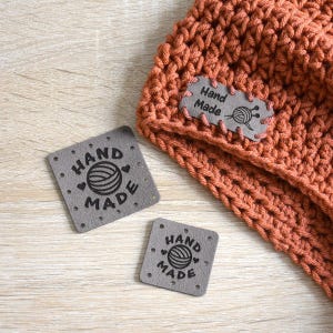 May include: A close-up of an orange crocheted beanie with a grey "Hand Made" label. Two additional grey labels with the text "Hand Made" and a yarn ball design are also visible. The labels are square with rounded corners.
