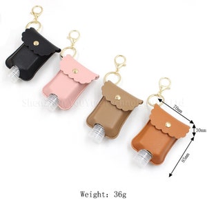 May include: Four keychain hand sanitizer holders in black, pink, brown, and orange. The holders are made of faux leather and have a scalloped edge. Each holder has a clear plastic bottle with a gold cap and a gold chain. The brown holder has measurements in millimeters.