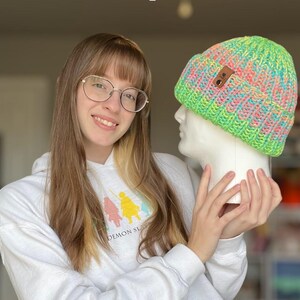 May include: A person wearing a white hoodie with the text "Demon Slayer" is holding a green, pink, and blue knit beanie with a brown leather tag. The beanie is on a white mannequin head.
