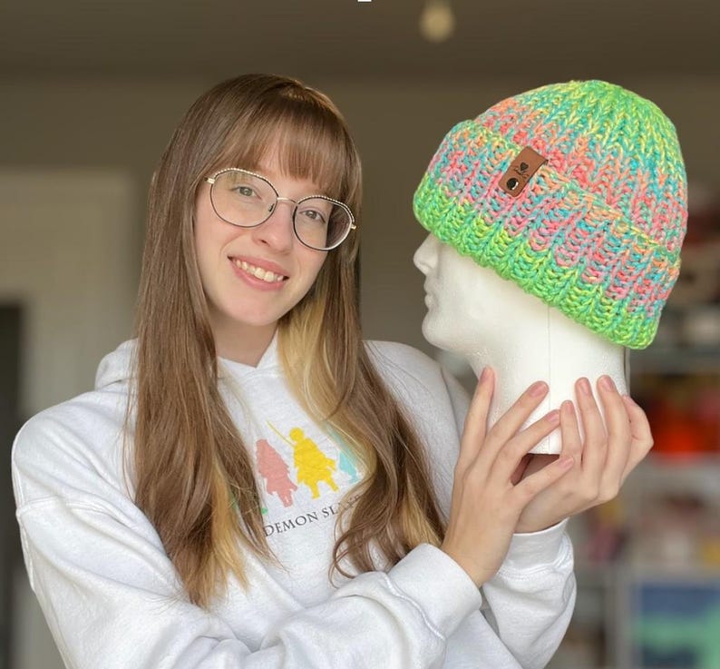 May include: A person wearing a white hoodie with the text "Demon Slayer" is holding a green, pink, and blue knit beanie with a brown leather tag. The beanie is on a white mannequin head.