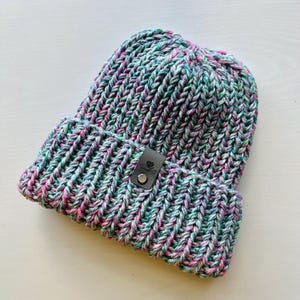 May include: A chunky knit beanie hat in a mix of pink, green, and white yarn. The hat has a black leather patch with a small metal button.