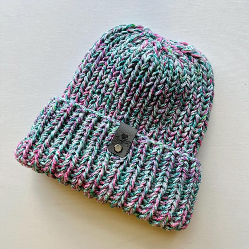 May include: A chunky knit beanie hat in a mix of pink, green, and white yarn. The hat has a black leather patch with a small metal button.