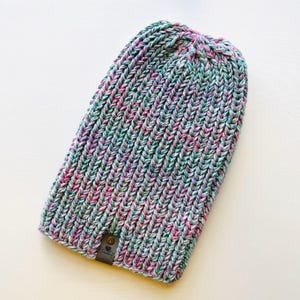 May include: A multi-colored knit beanie hat with a small leather tag. The hat is made of a soft, textured yarn in shades of pink, green, and white.
