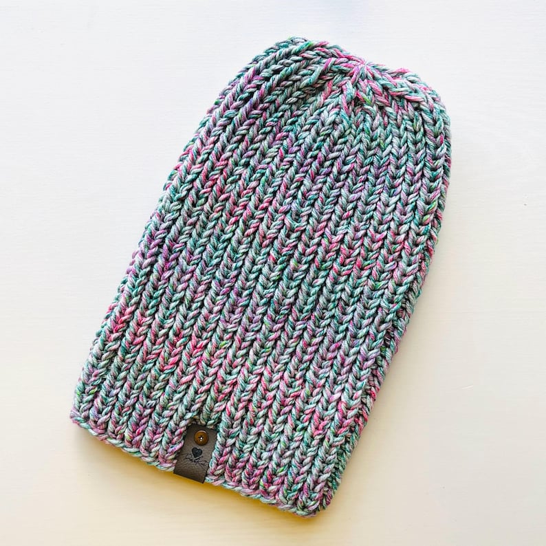 May include: A multi-colored knit beanie hat with a small leather tag. The hat is made of a soft, textured yarn in shades of pink, green, and white.