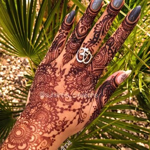 May include: A hand with intricate henna designs on the back of the hand and fingers. The nails are painted a light gray color. The person is wearing a silver ring with an Om symbol on the ring finger.