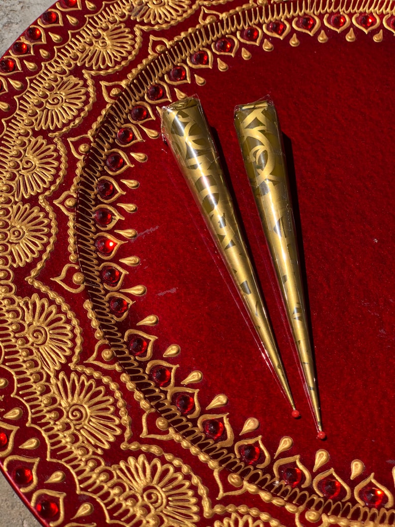 May include: Two gold henna cones on a red tray with gold and red decorative designs.