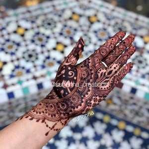 May include: Intricate henna design on a hand and forearm, featuring a large paisley pattern with intricate details. The design is dark brown and black, and the hand is resting on a tiled surface. The text "@suhenna_creations" and "suhenna" are visible on the image.