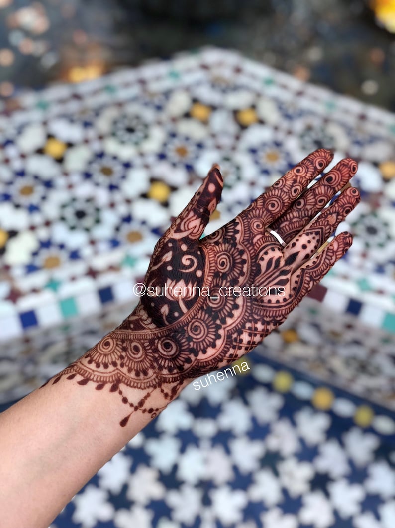 May include: Intricate henna design on a hand and forearm, featuring a large paisley pattern with intricate details. The design is dark brown and black, and the hand is resting on a tiled surface. The text "@suhenna_creations" and "suhenna" are visible on the image.