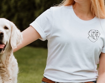 以下が含まれることがあります： 左胸に犬の頭の黒い輪郭が描かれた白いTシャツを着た女性。彼女はゴールデンレトリバーと一緒に草の上にひざまずいている。