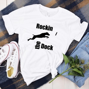 Op de afbeelding: Wit T-shirt met een zwart silhouet van een hond die over een stok springt met de tekst "Rockin' the Dock" erop gedrukt.