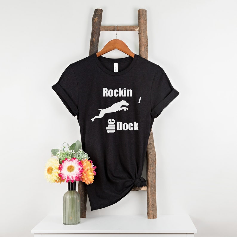 Op de afbeelding: Zwart T-shirt met een wit silhouet van een springende hond en de tekst "Rockin' the Dock".