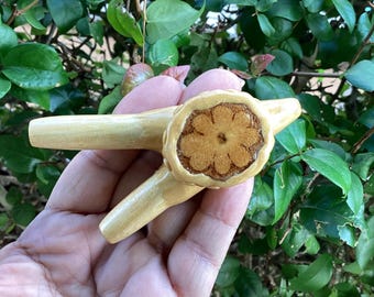 May include: A wooden pipe with a flower design carved into the bowl. The pipe is light brown and has two stems.