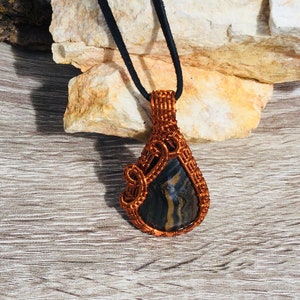 May include: A copper wire wrapped pendant featuring a black and gold striped stone. The pendant is hanging from a black cord.