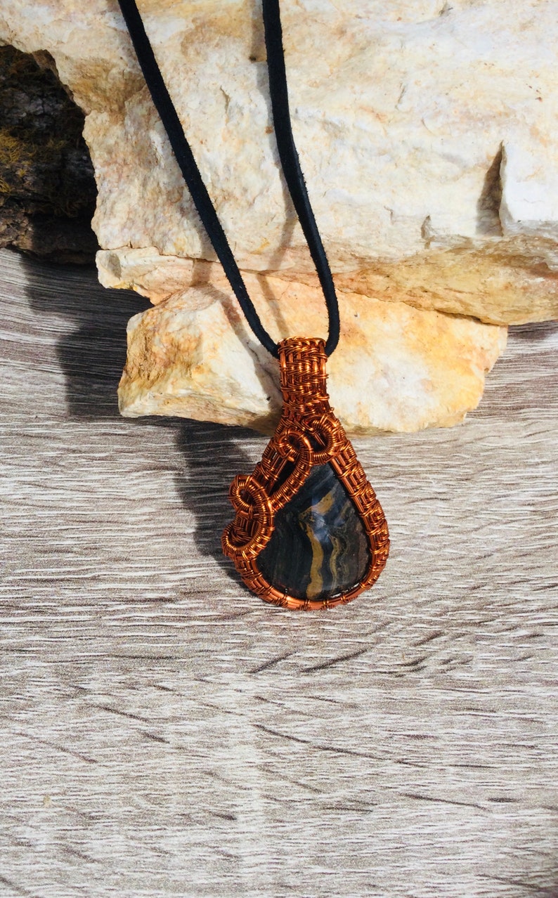 May include: A copper wire wrapped pendant featuring a black and gold striped stone. The pendant is hanging from a black cord.
