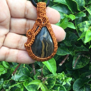 May include: A copper wire wrapped pendant featuring a black and gold striped stone. The pendant is teardrop shaped and hangs from a black cord.