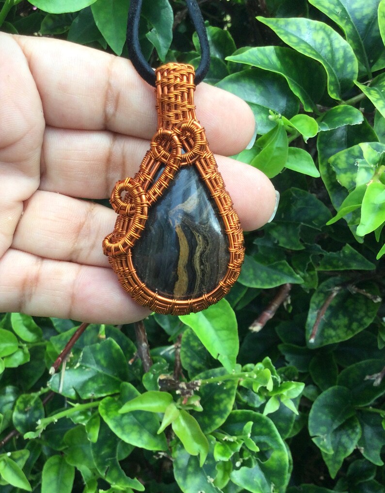 May include: A copper wire wrapped pendant featuring a black and gold striped stone. The pendant is teardrop shaped and hangs from a black cord.
