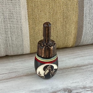 May include: A small, decorative bottle stopper with a carved wooden top and a black base. The base features a white and black mountain scene with colorful stripes. The stopper is approximately 3 inches tall.