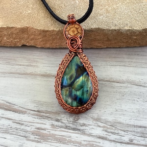 May include: A handcrafted copper wire-wrapped pendant featuring a teardrop-shaped labradorite stone. The stone displays iridescent blue, green, and yellow hues. The pendant hangs from a black cord necklace, showcasing the intricate wirework and natural stone.
