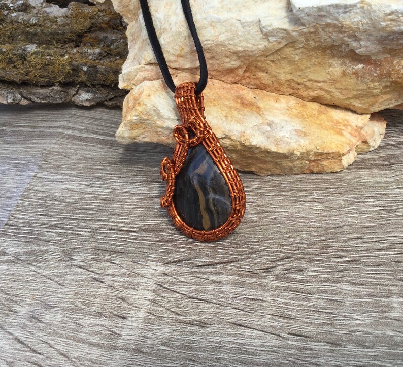 May include: A teardrop-shaped pendant with a black and gold stone wrapped in copper wire. The pendant is hanging from a black cord.