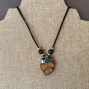 Può includere: Un ciondolo in legno marrone con un'incisione di pietra verde, su un cordoncino nero con perline argentate e marroni. Il ciondolo ha la forma di un quadrifoglio.