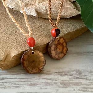 May include: Two handmade necklaces with natural wood pendants and red beads. The pendants are round, with a unique wood grain pattern. The necklaces have beige macrame cords. The background features a stone surface and a wooden surface.
