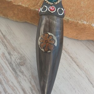 May include: A black horn pendant with a silver and gold floral design, accented with red and green stones. The pendant is attached to a black cord.
