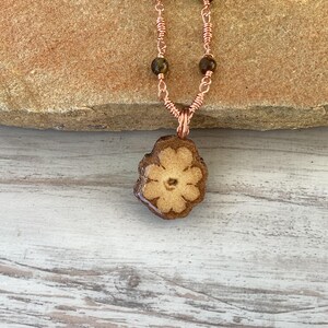 May include: A copper chain necklace with a brown flower-shaped pendant. The pendant is made of a natural material with a light brown center and a darker brown outer edge. The chain has small brown beads.