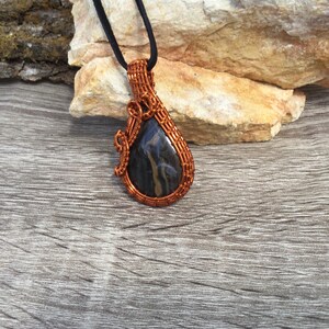 May include: A copper wire wrapped pendant featuring a black and gold stone. The pendant is hanging from a black cord.