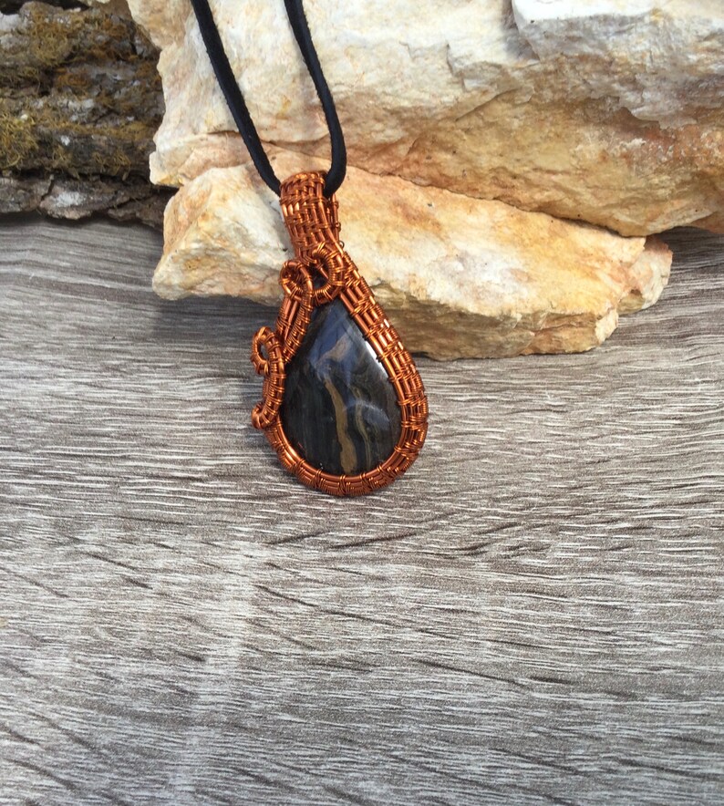 May include: A copper wire wrapped pendant featuring a black and gold stone. The pendant is hanging from a black cord.