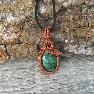 May include: A green gemstone pendant wrapped in copper wire. The pendant is hanging from a black cord.