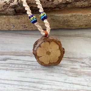 May include: A handmade necklace featuring a pendant made from a natural, light brown material with a floral design. The pendant is suspended from a cream-colored, braided cord, accented with blue and yellow beads. The necklace is displayed on a wooden surface.