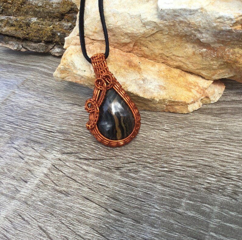 May include: A teardrop-shaped pendant with a black and silver stone wrapped in copper wire. The pendant is hanging from a black cord.