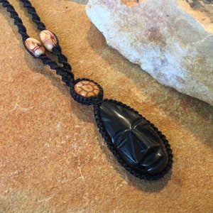 May include: Black wooden pendant necklace with a carved face design. The necklace is made with black cord and brown wooden beads.