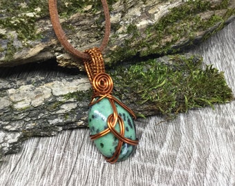 May include: A green and black speckled stone pendant wrapped in copper wire. The pendant is hanging from a brown leather cord.