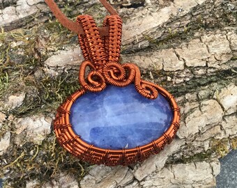 May include: A copper wire wrapped pendant featuring a light blue oval gemstone. The pendant is attached to a brown leather cord.