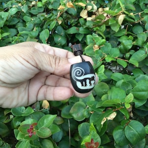 Puede incluir: Collar con colgante negro con diseño de mono tallado. El colgante es de color oscuro con una silueta de mono de color claro y un diseño en espiral. El collar se sostiene frente a un fondo de hojas verdes.