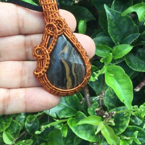 May include: A teardrop-shaped pendant with a black and gold stone wrapped in copper wire. The wire is woven into a decorative pattern.