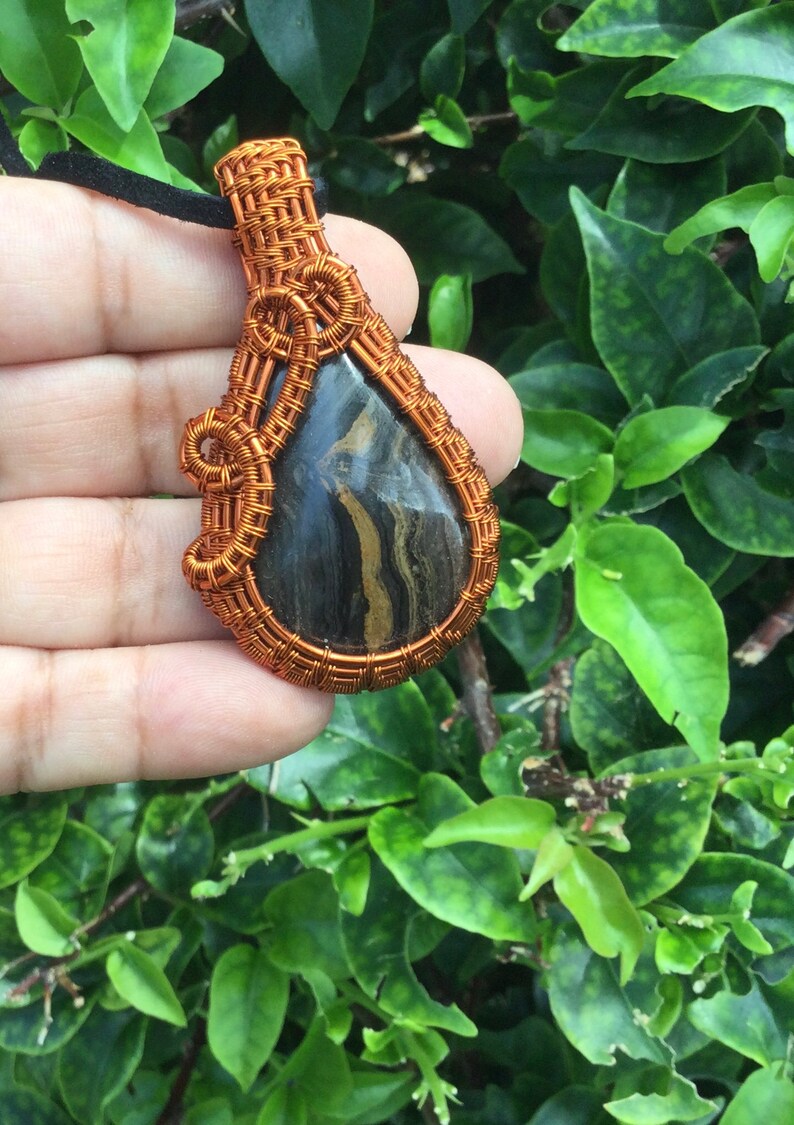May include: A teardrop-shaped pendant with a black and gold stone wrapped in copper wire. The wire is woven into a decorative pattern.