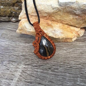 May include: A copper wire wrapped pendant featuring a black and silver stone. The pendant is hanging from a black cord.