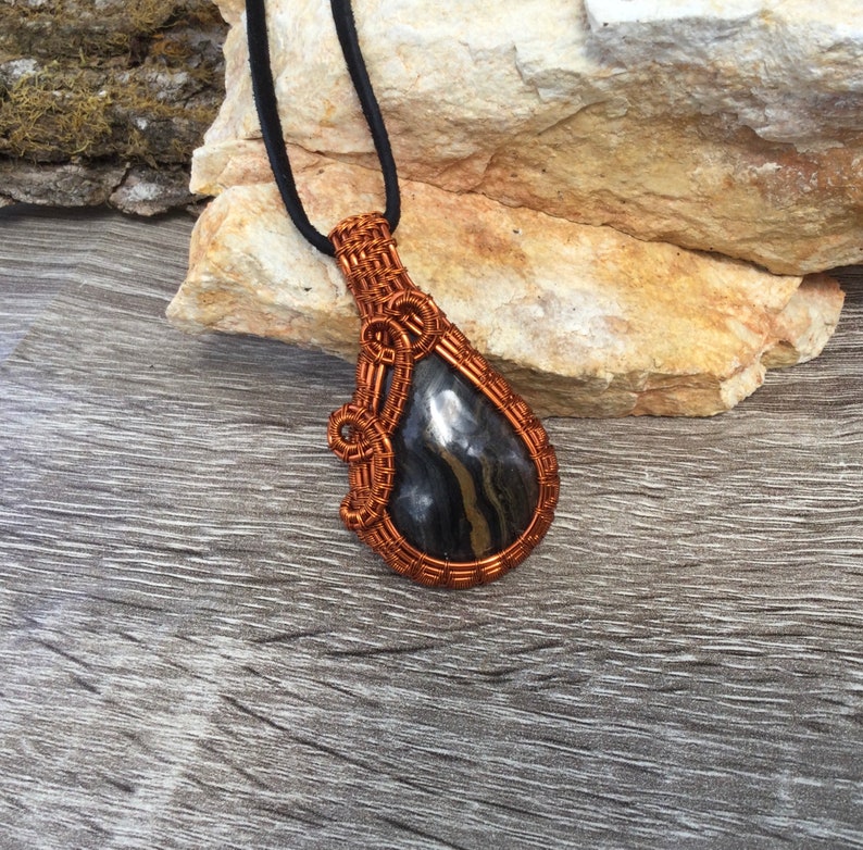 May include: A copper wire wrapped pendant featuring a black and silver stone. The pendant is hanging from a black cord.