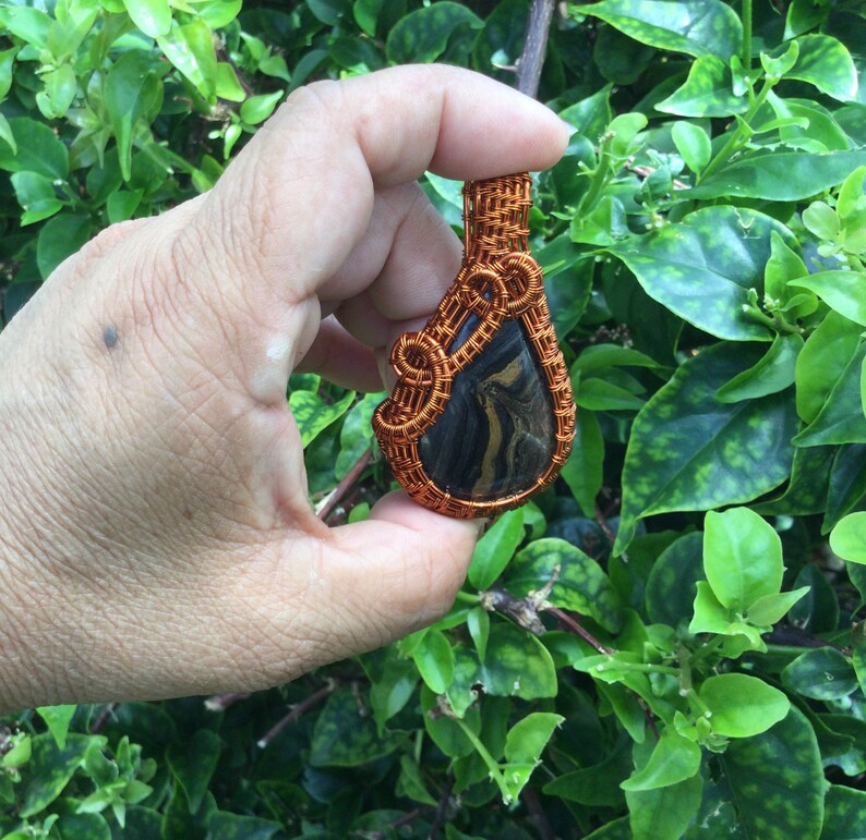 May include: A copper wire wrapped pendant featuring a black and gold stone. The pendant is teardrop shaped and has a woven wire design.