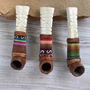 May include: Three white wooden pipes with brown leather and colorful woven fabric accents. The pipes are carved with geometric designs.