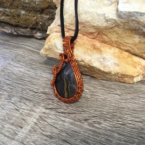 May include: A copper wire wrapped pendant featuring a black and brown agate stone. The pendant is hanging from a black cord.