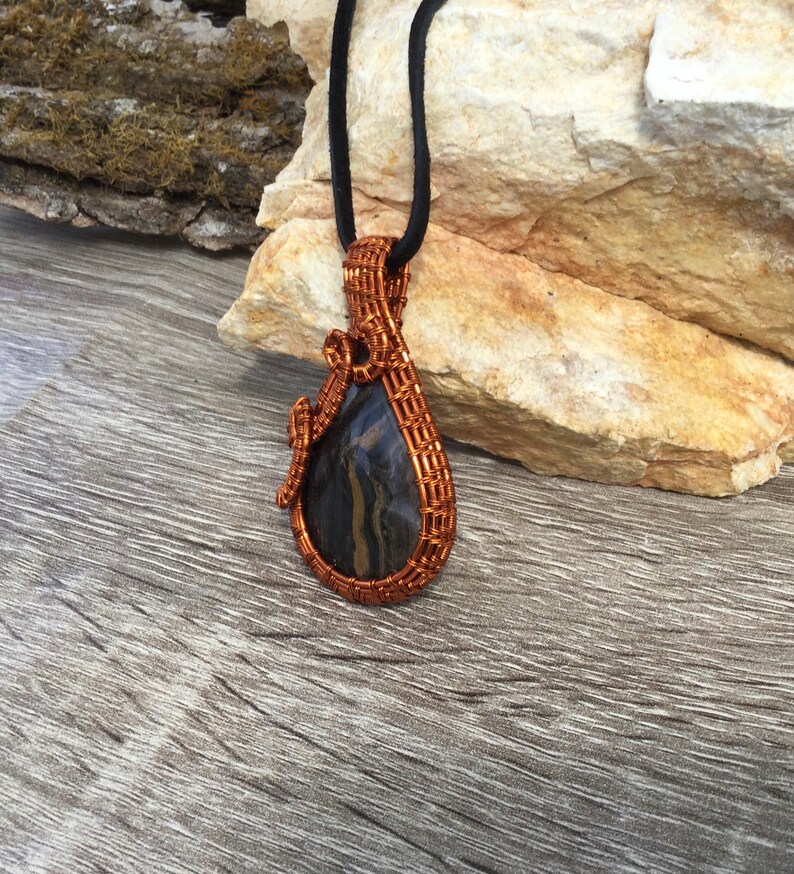 May include: A copper wire wrapped pendant featuring a black and brown agate stone. The pendant is hanging from a black cord.