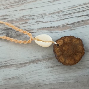 May include: A beige cord necklace with a white cowrie shell and a brown wooden pendant with a circular design.