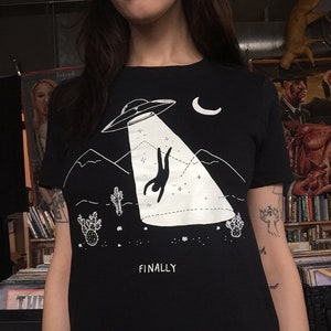 May include: Black t-shirt with a white graphic of a person falling from a UFO with the text "FINALLY" underneath.